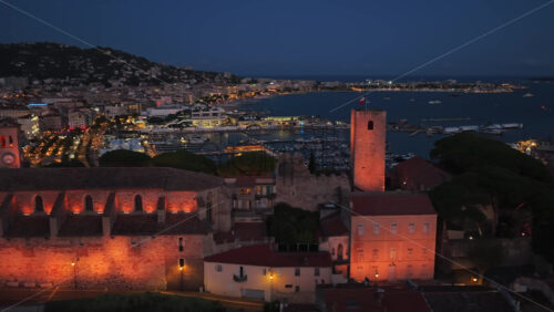 Video - Aerial drone view of Cannes Old Town during twilight, with the illuminated Church of Our Lady of Esperance and the medieval watchtower overlooking the yacht filled harbor and the Bay of Cannes
