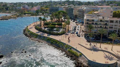 Video - Aerial drone view of Antibes seafront promenade, curved walkway lined with palm trees, overlooking the Mediterranean Sea and modern city buildings