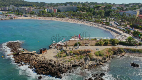 Video - Aerial drone view of a beach and coastline in Antibes, France, with turquoise waters, rocky breakwaters, beach clubs, and sunbathers enjoying the summer atmosphere