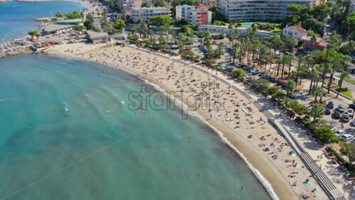 Video - Aerial drone view of Antibes beach and promenade, lined with palm trees and apartments, with turquoise Mediterranean waters and swimmers in the bay