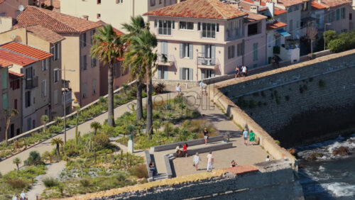 Video - Antibes, France - September 4, 2025: Aerial drone view of people walking along the historic stone ramparts of the Old Town, with cactus plants, palm trees, and Mediterranean landscaping