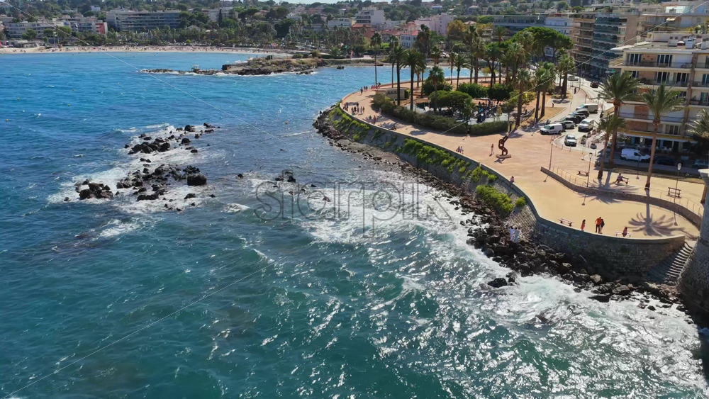 Video - Aerial drone view of waves crashing against the rocky shoreline of Antibes, with the seafront promenade and palm trees visible above