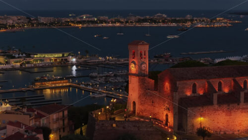 Video - Aerial drone view of Cannes Old Town during twilight, with the illuminated Church of Our Lady of Esperance and the medieval watchtower overlooking the yacht filled harbor and the Bay of Cannes