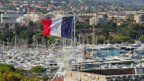 Video - Aerial drone view of the French flag waving above Antibes marina, with luxury yachts, sailboats, and a Ferris wheel in the background