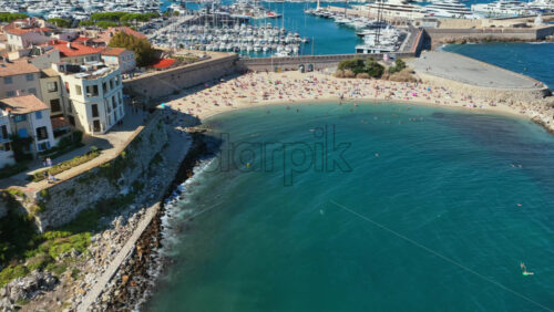 Video - Aerial drone view of Antibes sandy beach filled with sunbathers, turquoise waters, and the nearby marina with luxury yachts