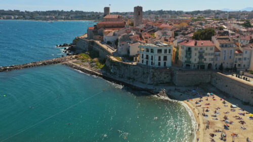 Video - Aerial drone view of Antibes Old Town, medieval ramparts, sandy beach, and marina with yachts, seen from above the sea