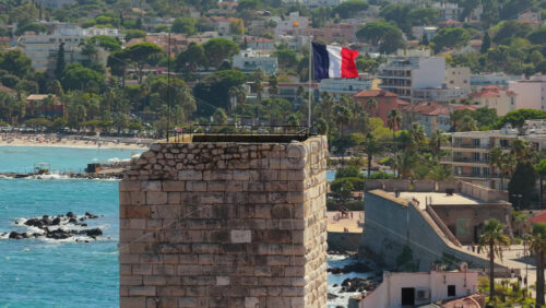 Video - Aerial drone view of the French flag waving above Antibes Old Town medieval tower, with palm-lined promenade and the Mediterranean Sea in the background