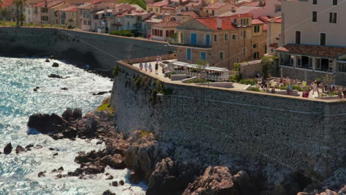 Video - Aerial drone view of Antibes stone ramparts and seaside promenade with visitors walking along the walls, overlooking the rocky Mediterranean coast
