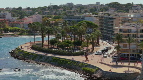 Video - Aerial drone view of a coastal promenade lined with palm trees and public art sculptures, with people strolling near the Mediterranean Sea, Antibes, France