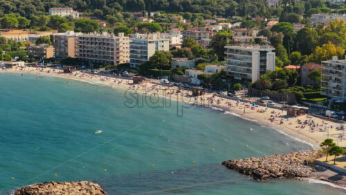 Video - Aerial drone view of Antibes sandy beach filled with sunbathers and swimmers, with seafront apartment buildings and green hills in the background