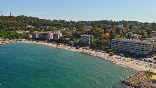 Video - Aerial drone view of Antibes sandy beach filled with sunbathers and swimmers, with seafront apartment buildings and green hills in the background
