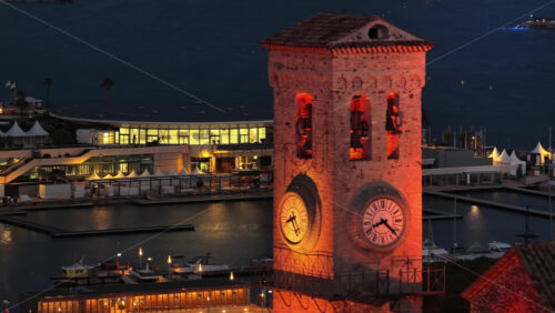 Video - Aerial drone view of the historic Cannes clock tower at dusk, with the bustling city center in the background, glowing with evening lights
