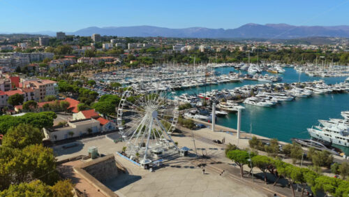 Video - Aerial drone view of the Ferris wheel in Antibes near the marina, with hundreds of yachts docked in the port and the mountains in the background