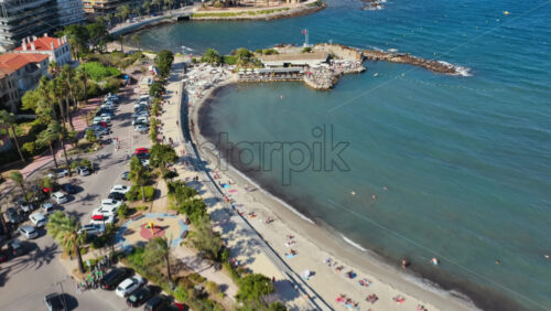 Video - Aerial drone view of Nice's sandy beach filled with sunbathers and swimmers, with palm lined Promenade des Anglais and seafront buildings behind