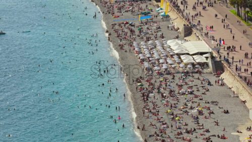 Video - Nice, France - September 5, 2025: Aerial drone view of crowded Nice beach with people sunbathing on pebbles and swimming in turquoise Mediterranean waters