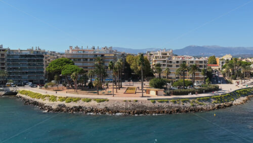 Video - Aerial drone view of the Antibes seafront promenade lined with palm trees and modern apartments, with the Mediterranean Sea and distant mountains in the background