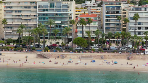 Video - Cannes, France - September 6, 2025: Aerial drone view of the city beach with people relaxing on the sand and swimming in the turquoise sea, surrounded by palm trees and city buildings