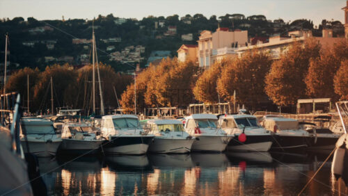Video - Row of boats docked near autumn trees and city buildings, reflecting golden evening light on calm water