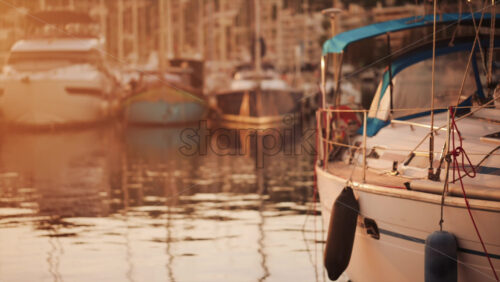 Video - Elegant sailboats lined up in a marina during golden hour, calm water reflecting the evening sun