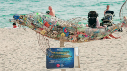 Video - Cannes, France - October 4, 2025: Metal dolphin sculpture filled with plastic waste displayed on a sandy beach by the sea, raising awareness about ocean pollution