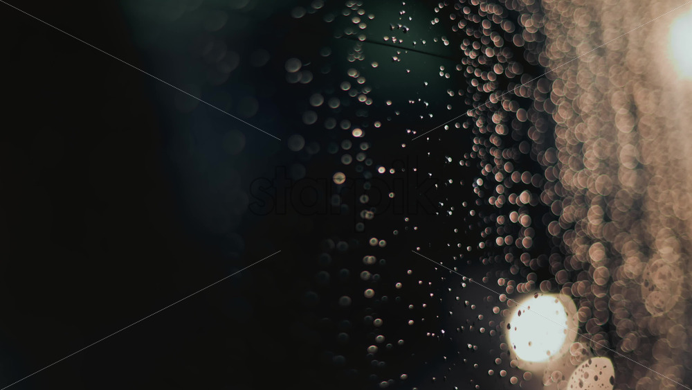 Video - Close up of raindrops on a glass window, softly illuminated by blurred bokeh lights in the background