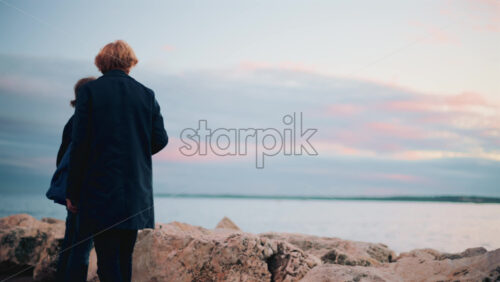 Video - Cannes, France - October 20, 2025: Two elderly people standing on coastal rocks, gazing at the calm sea under a pastel sunset sky