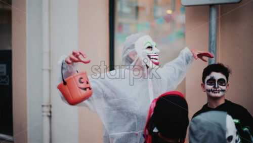 Video - Cannes, France - October 31, 2025: A person wearing a wide grinning clown mask, raises their hands playfully, holding an orange pumpkin shaped bucket near children in skeleton costumes on Halloween