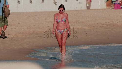 Video - Cannes, France - October 4, 2025: Woman in a colorful bikini walking along the sandy beach at sunset