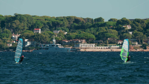 Video - Cannes, France - October 4, 2025: Two windsurfers gliding across blue sea with lush green coastline and luxury villas in the background