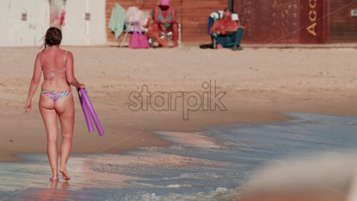 Video - Cannes, France - October 4, 2025: Rear view of a woman in a colorful bikini walking away along the sandy shore carrying a purple pool float