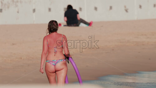 Video - Rear view of a woman in a colorful bikini walking away along the sandy shore carrying a purple pool float