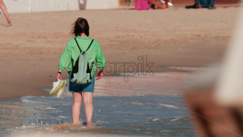 Video - Young woman with backpack walking barefoot on the beach holding her shoes