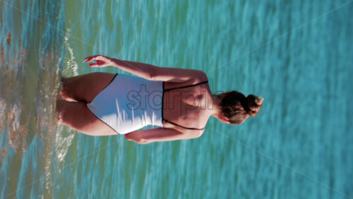 Video - A young woman walks slowly into the calm turquoise water on a sunny summer day in Cannes, France. Vertical