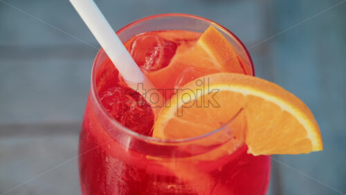Video - Close up of a refreshing red orange cocktail with orange slice and ice cubes, captured outdoors on a sunny day in Cannes