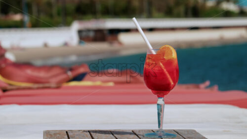 Video - A bright orange red orange cocktail placed on a wooden table at a beach lounge with the blue sea and sunbathers in the background