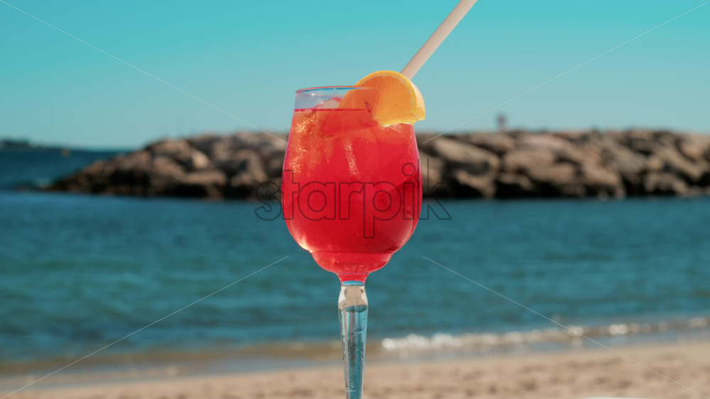 Video - Refreshing red orange cocktail with ice and orange slice against the deep blue Mediterranean Sea