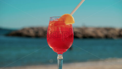 Video - Refreshing red orange cocktail with ice and orange slice against the deep blue Mediterranean Sea