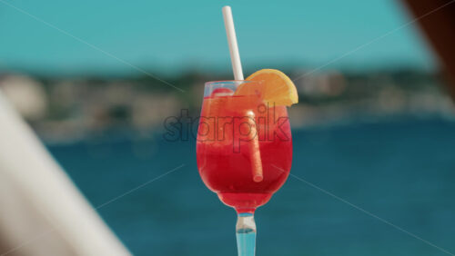Video - Refreshing red orange cocktail with ice and orange slice against the deep blue Mediterranean Sea