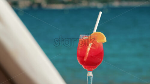 Video - Refreshing red orange cocktail with ice and orange slice against the deep blue Mediterranean Sea