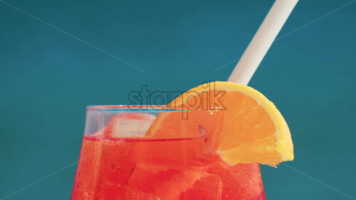 Video - Close up of a refreshing red orange cocktail with orange slice and ice cubes, captured outdoors on a sunny day in Cannes
