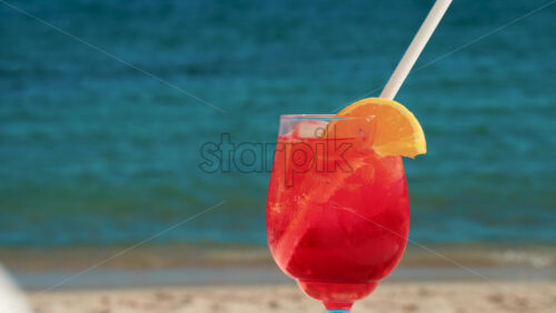 Video - Refreshing red orange cocktail with ice and orange slice against the deep blue Mediterranean Sea