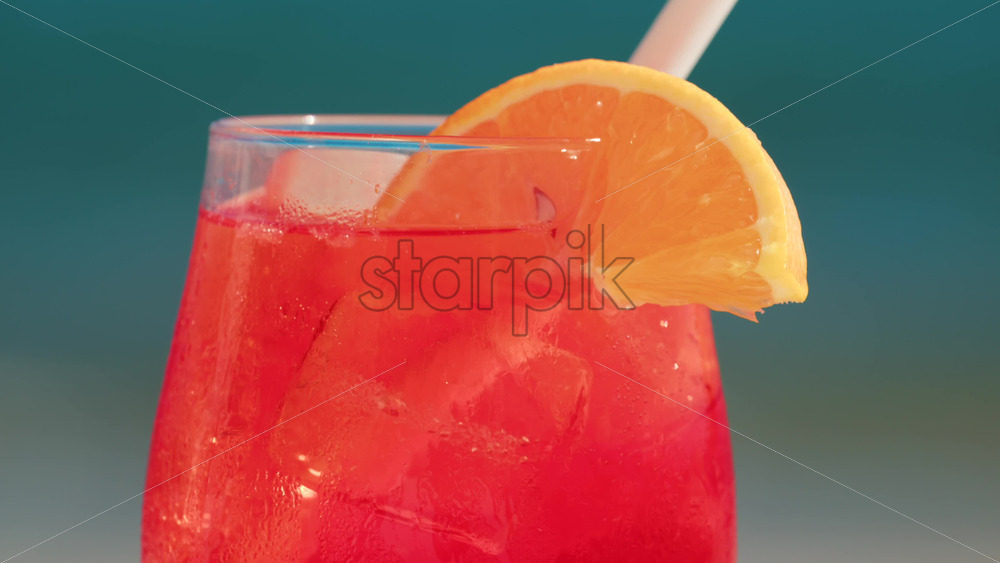 Video - Close up of a refreshing red orange cocktail with orange slice and ice cubes, captured outdoors on a sunny day in Cannes