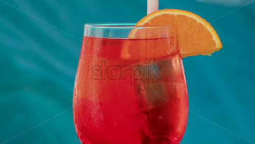 Video - A hand stirs a bright red orange with an orange slice against a turquoise background