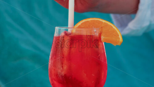 Video - A hand stirs a bright red orange with an orange slice against a turquoise background