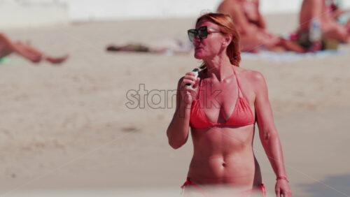 Video - Cannes, France - October 6, 2025: A woman in a red bikini stands on the sandy beach vaping and relaxing by the sea