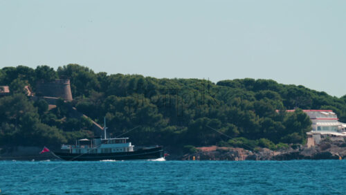 Video - A stylish black yacht sails along the Cannes coastline surrounded by pine trees and villas under clear blue skies on the French Riviera