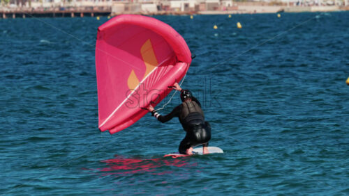 Video - A man in a wetsuit practices wing foiling with a bright pink sail on the blue Mediterranean waters of Cannes, France
