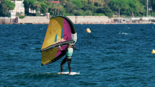 Video - Cannes, France - October 6, 2025: A woman balances on a wing foil board holding a colorful yellow and pink sail on a sunny day
