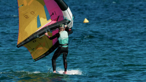 Video - Cannes, France - October 6, 2025: A woman balances on a wing foil board holding a colorful yellow and pink sail on a sunny day