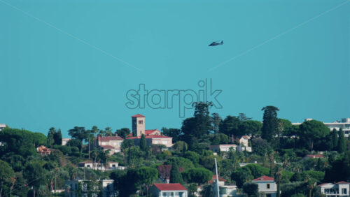 Video - A black helicopter flies above the green hills and luxury villas of Cannes, France, under a clear blue sky